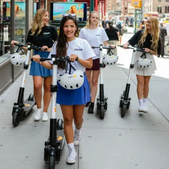Four riders walking Ride by iPakket e-scooters along a city sidewalk